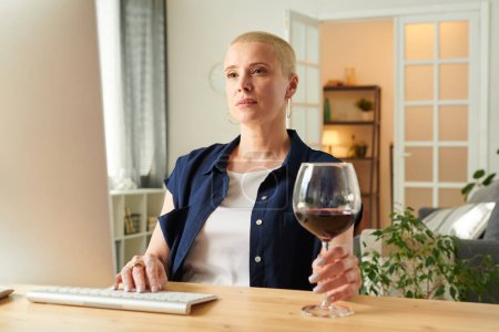 Téléchargez les photos : Belle femme assise à table avec un verre de vin rouge et utilisant un ordinateur dans le salon - en image libre de droit
