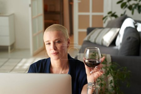 Photo for Serious pretty woman enjoying glass of red wine while sitting at table in front of monitor and watching video - Royalty Free Image