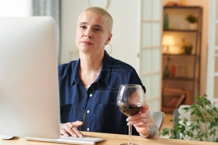 Mujer bonita con el pelo corto que se comunica en línea usando el ordenador en la mesa en casa y beber una copa de vino tinto