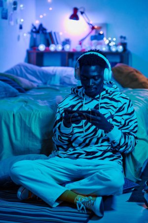 Photo for African teenage boy in wireless headphones sitting on floor in dark bedroom and playing video game on smartphone - Royalty Free Image