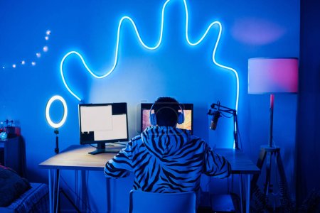 Photo for Rear view of young gamer in headphones playing online video games while streaming on social media in dark neon room - Royalty Free Image