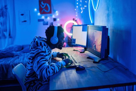 Photo for African gamer boy in wireless headphones typing on keyboard while sitting at table in front of computer and playing video game - Royalty Free Image