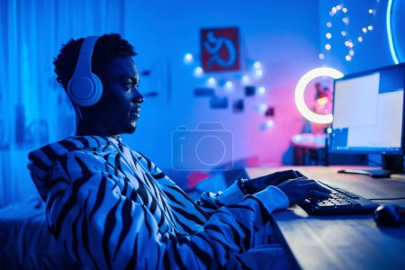 Téléchargez les photos : Arican adolescent garçon dans les écouteurs sans fil jouer jeu vidéo sur ordinateur tout en étant assis à la table dans la chambre noire - en image libre de droit