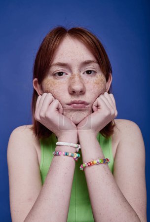 Téléchargez les photos : Portrait d'adolescente aux cheveux roux avec des bracelets faits à la main sur ses bras en regardant la caméra sur fond bleu - en image libre de droit