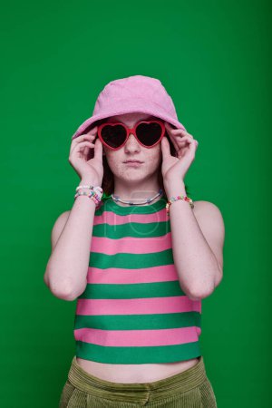 Téléchargez les photos : Portrait d'adolescente en lunettes de soleil et chapeau panama debout sur fond vert - en image libre de droit
