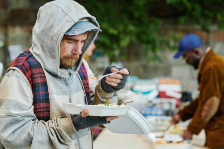 Foto de Hombre sin hogar con ropa rota caliente comiendo comida al aire libre dando por voluntarios durante la caridad - Imagen libre de derechos
