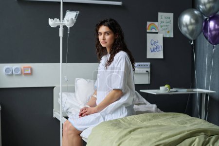 Foto de Retrato de mujer joven con gotero mirando a la cámara mientras está sentada en la cama en la sala decorada con globos - Imagen libre de derechos