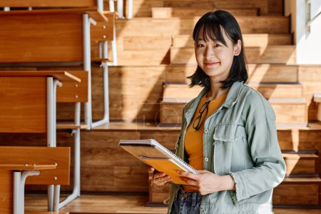 Photo for Portrait of Asian student with textbooks smiling at camera while standing at auditorium of university - Royalty Free Image