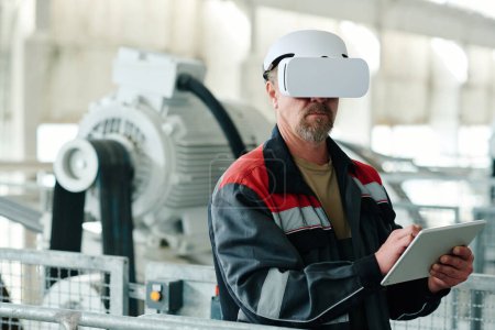 Photo for Mature engineer in uniform using digital tablet to watch production of material with VR glasses - Royalty Free Image
