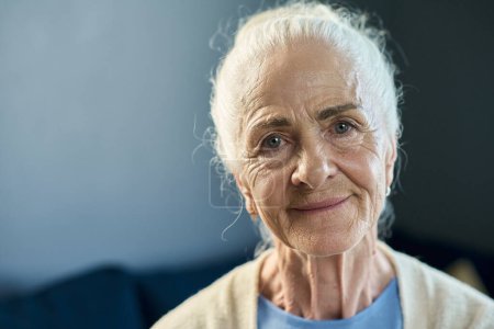 Foto de Rostro de mujer anciana sonriente con el pelo blanco mirándote con sonrisa mientras está sentada frente a la cámara sobre fondo azul en aislamiento - Imagen libre de derechos