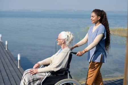 Vue latérale de la jeune petite-fille multiethnique en casual poussant fauteuil roulant avec grand-mère tout en marchant le long de la jetée au bord de l'eau