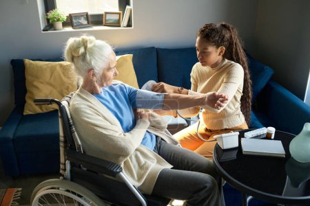 Téléchargez les photos : Adolescente multiethnique mettant la manche sur le bras de la grand-mère avant de mesurer la pression artérielle alors qu'elle est assise devant elle dans le salon - en image libre de droit