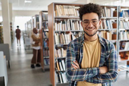 Téléchargez les photos : Portrait de l'étudiant en lunettes souriant à la caméra tout en se tenant dans la bibliothèque du collège - en image libre de droit