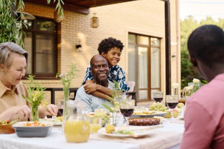 Photo for Cheerful African American boy in Hawaiian shirt embracing his grandfather by dinner while standing close to him and looking at his parents - Royalty Free Image