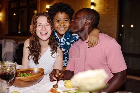 Photo for Young brunette woman in white dress laughing while cute boy embracing his happy intercultural parents sitting by served table during dinner - Royalty Free Image