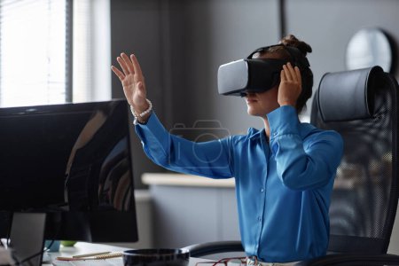 Photo for Young woman testing new program in VR glasses while sitting at her workplace - Royalty Free Image