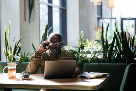 Téléchargez les photos : Homme âgé utilisant un ordinateur portable et parlant au téléphone alors qu'il était assis dans un café - en image libre de droit