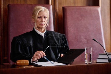 Juez femenina rubia madura seria de la corte suprema de justicia en manto negro sentado en silla de cuero carmesí por escritorio de madera en la sala de audiencias