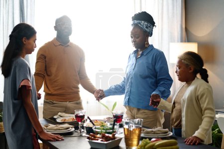 Téléchargez les photos : Jeune famille de quatre personnes tenant par la main autour d'une table servie avec de la nourriture et des boissons maison préparées pour le dîner de Thanksgiving - en image libre de droit
