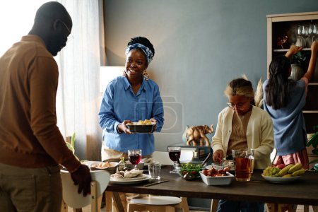 Fröhliche junge Afroamerikanerin mit Tablett mit gekochtem Essen blickt ihren Mann an und serviert festlichen Tisch mit hausgemachten Speisen und Getränken