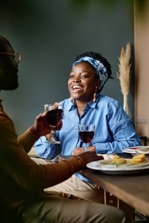 Téléchargez les photos : Jeune femme joyeuse regardant son mari avec un verre de vin rouge fait maison assis devant elle par table de fête servie - en image libre de droit