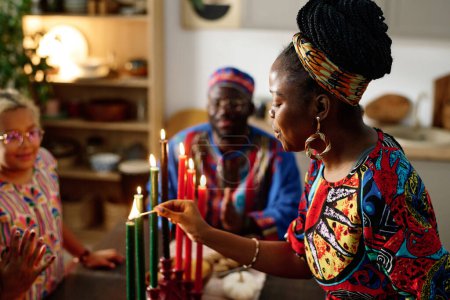 Téléchargez les photos : Vue latérale de la jeune femme afro-américaine en vêtements ethniques brûlant des bougies symbolisant sept principes principaux de la culture nationale - en image libre de droit