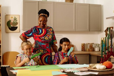 Téléchargez les photos : Jeune femme afro-américaine en tenue nationale regardant ses filles couper du papier tout en préparant des cartes de vœux pour la célébration de la kwanzaa - en image libre de droit