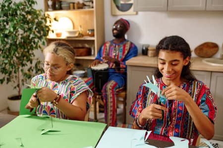 Téléchargez les photos : Filles créatives en tenue ethnique couper les mains de papier vert et bleu tout en étant assis à table et faire bricolage artisanat pour la célébration kwanzaa - en image libre de droit