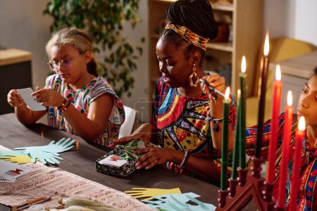 Téléchargez les photos : Focus sur la jeune femme en costume national emballage cadeaux kwanzaa pour sa famille tout en étant assis à côté de sa fille brûler sept bougies - en image libre de droit