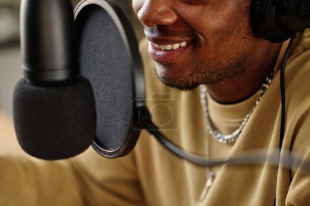 Téléchargez les photos : Sourire de jeune hôte afro-américain parlant au microphone tout en faisant un podcast ou une interview avec un invité pour le public en ligne - en image libre de droit