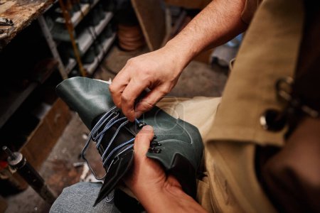 Téléchargez les photos : Mains de cordonnier professionnel masculin mettant lacet dans la partie supérieure de la botte en cuir noir non fini tout en étant assis dans l'atelier - en image libre de droit