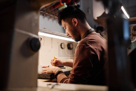 Téléchargez les photos : Vue latérale du jeune cordonnier asiatique travaillant sur le nouvel ordre des clients de son atelier de travail du cuir assis par le lieu de travail - en image libre de droit