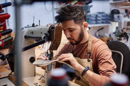 Téléchargez les photos : Jeune homme asiatique en vêtements de travail assis devant la machine à coudre dans l'atelier et de mettre ensemble des détails de nouvelles chaussures - en image libre de droit