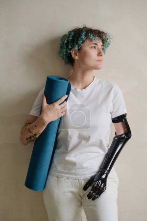 Photo for Portrait of pensive girl with prosthetic arm holding yoga mat and looking away - Royalty Free Image