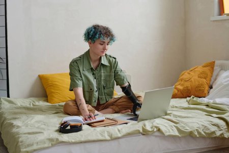 Photo for College student with prosthetic arm sitting on bed and doing homework - Royalty Free Image