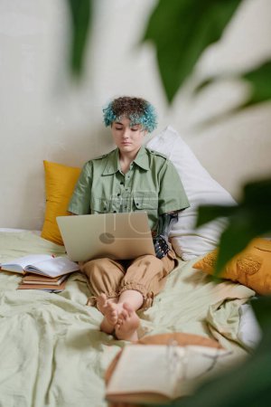 Photo for Smart girl with prosthetic arm sitting on bed and working on laptop - Royalty Free Image