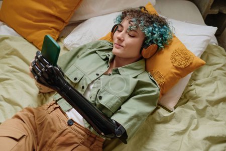 Foto de Mujer joven sonriente con brazo protésico escuchando música en los auriculares y revisando las redes sociales en el teléfono inteligente - Imagen libre de derechos