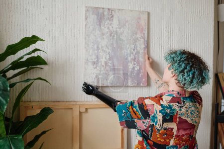 Photo for Artist with prosthetic arm hanging picture on wall in her apartment - Royalty Free Image