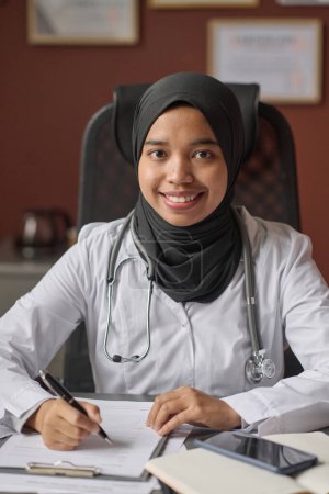 Foto de Doctora joven en hijab sentada en el escritorio y sonriendo a la cámara mientras rellena documentos - Imagen libre de derechos