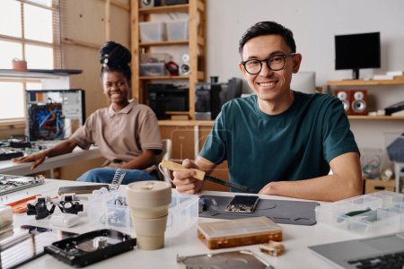 Photo for Caucasian technician smiling at camera while soldering cellphone, his black coworker sitting on background - Royalty Free Image