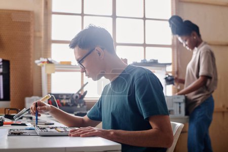 Photo for Young man repairing PC element with soldering iron, his black coworker standing on background - Royalty Free Image