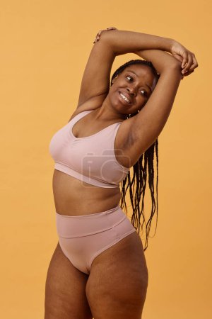 Vertikales mittellanges Studioporträt einer lebenslustigen Afroamerikanerin, die bequeme Unterwäsche trägt und für die Kamera posiert, warmer apricotfarbener Hintergrund