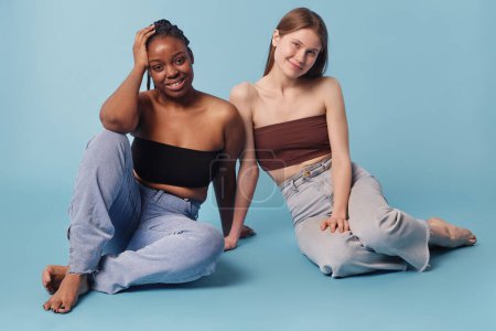Studioporträt zweier junger, ethnisch unterschiedlicher Frauen in Bandeau-Tops und Jeans, die auf dem Boden sitzen und in die Kamera schauen, babyblauer Hintergrund