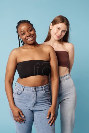 Vertical medium long studio portrait of two young ethnically diverse women with no makeup wearing bandeau tops and jeans posing for camera