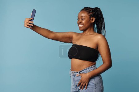 Mittleres Studioporträt einer fröhlichen jungen Afroamerikanerin mit Bandeau-Oberteil und Jeans, die ein Selfie mit dem Smartphone macht