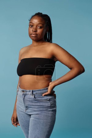Vertical medium long portrait of young African American woman wearing bandeau top and jeans posing for camera in studio