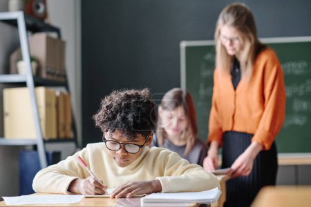 Selective focus shot of African American teen boy writing in notebook during lesson, young teacher helping girl with task in background