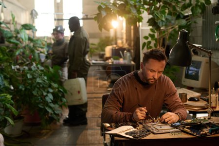 Téléchargez les photos : Mise au point sélective de l'homme caucasien réparer vieil ordinateur tandis que ses amis prenant soin des plantes dans un abri post-apocalyptique - en image libre de droit