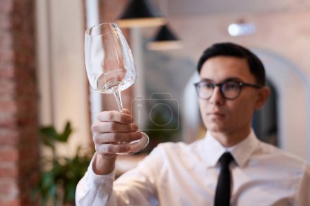 Selective focus medium closeup of young Asian restaurant manager holding wine glass checking its cleanliness