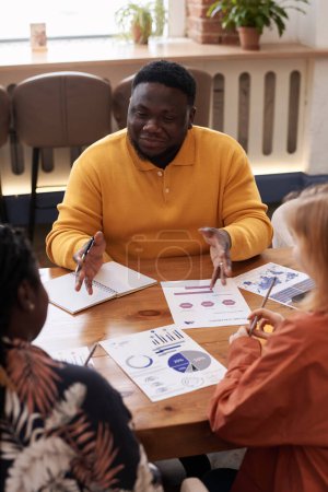 Vertical high angle over-the-shoulder of young Black man and two diverse women wearing casual clothes having business meeting in modern restaurant